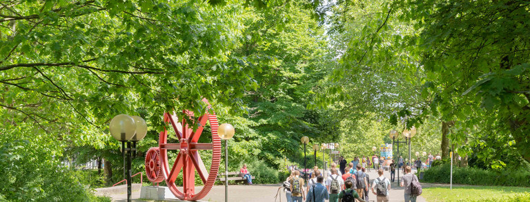 Studierende sind im Sommer auf dem Campus Nord, rote Zahnräder im Hintergrund.